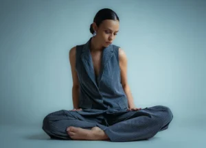 A woman in a vest and pants sits cross-legged.
