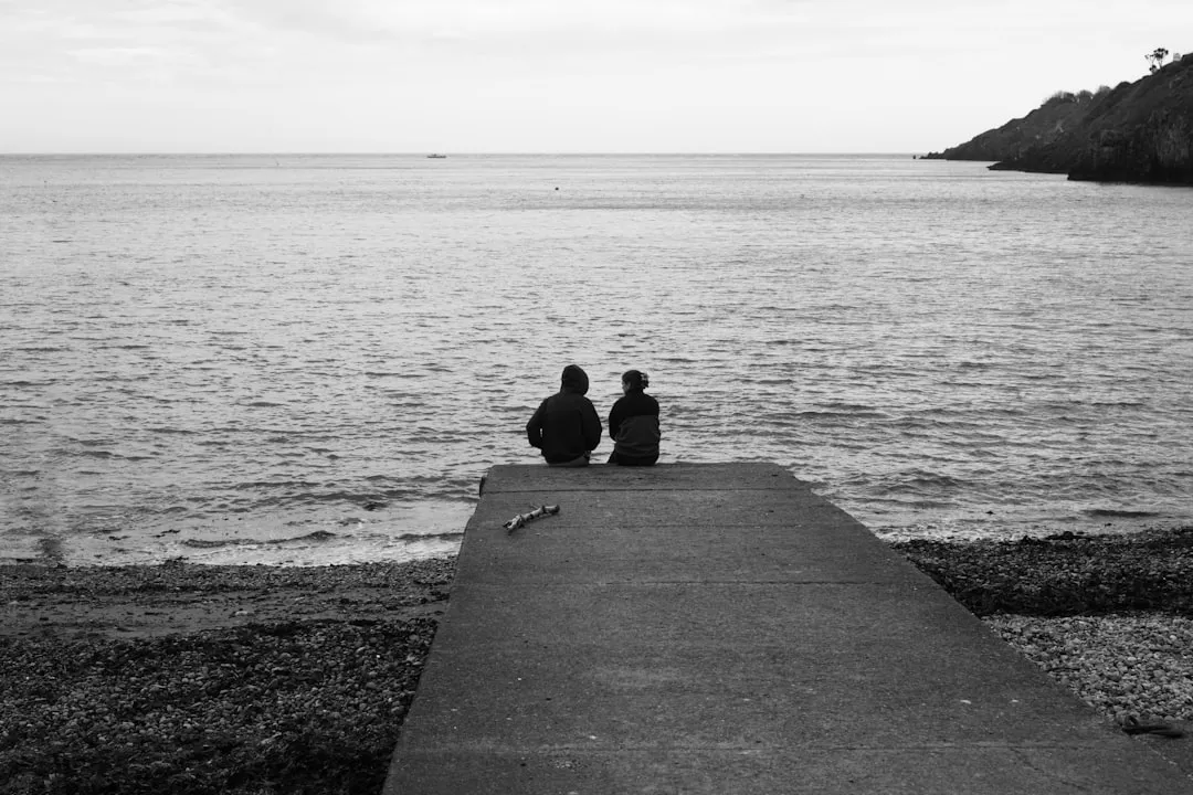 Two people sit on a pier overlooking the ocean.
