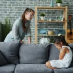 Mother scolds crying daughter on sofa in living room.